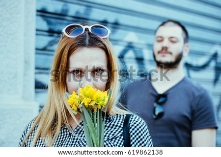 Photo of woman smelling the bouquet of flowers in front of burred man with sunglasses - steel street garage roller door with graffiti in background. Young couple enjoying hot spring day