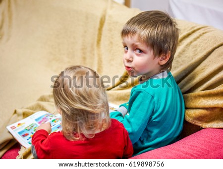 brother teach sister. two small kids brother and sister sit on burgundy sofa and reading book with pictures. Older brother show to small blonde sister picture on soft book in homely atmosphere