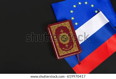 Small flag of EU and Russia and a holly book Koran on a black plastic surface. Translation from Arabic- the book contains verses from  Koran