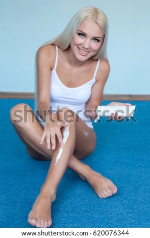 Luxury beautiful blonde girl or woman with blue eyes smear white cream or lotion on body. Body cosmetology procedure with cream. Smiling girl in white shirt. Body care, legs care. Smear cream