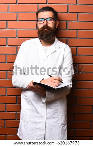 Bearded man, long beard. Brutal caucasian doctor or unshaven hipster, postgraduate student holding notebook with pen in medical gown and glasses on brown brick wall studio background. Medicine concept