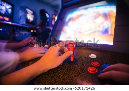 POV angle on hands playing vintage arcade game with an opponent, in a dark shade room full of arcade games and pinballs Royalty-Free Stock Photo #620171426