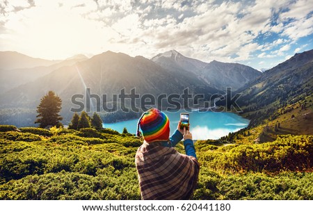 Tourist woman in rainbow hat and brown poncho taking picture with her smartphone  in beautiful Mountain Lake in Kazakhstan