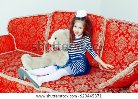 Little girl in blue striped dress and a white bow on her head.Girl sitting on sofa with Teddy bear.Creative toning of a photograph.
