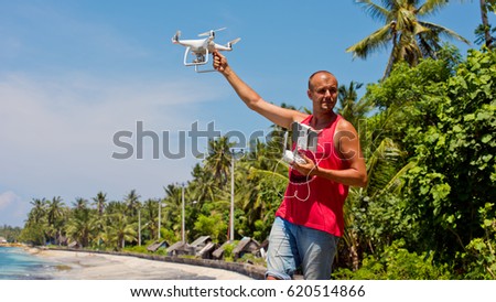 A young man lands a white drone in his hand in background of a tropical beach.
