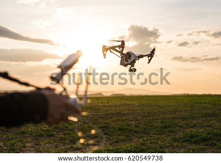 Man controls quadrocopter flight. Flying the copter over a field over sunset sky. Remote control in a man's hands.