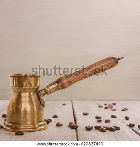 A toned square photo of a vintage coffee pot with a wooden handle, side view on a light wooden background texture with copyspace, with coffee beans scattered around