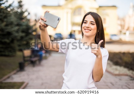 Young happy caucasian woman wearing casual clothes is taking a selfie with her smartphone at the park during a sunny day