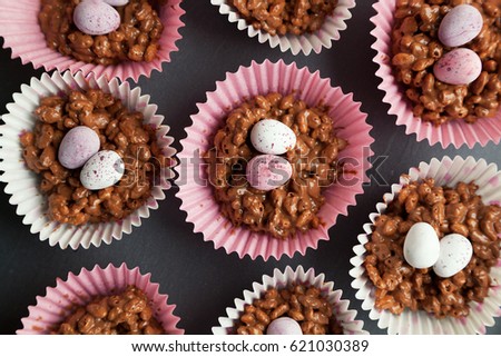 Close up of rice krispie cakes in pink and white cup cake cases with sugar coated eggs on top on a black slate background. Royalty-Free Stock Photo #621030389