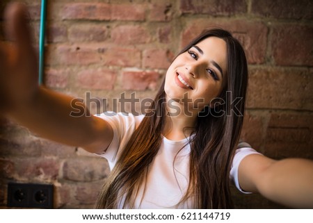 Beautiful young woman making self-portrait on a smartphone on a brick wall background