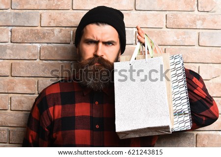Bearded man, long beard. Brutal caucasian serious hipster with moustache holding shopping packages in red black checkered shirt on beige brick wall studio background