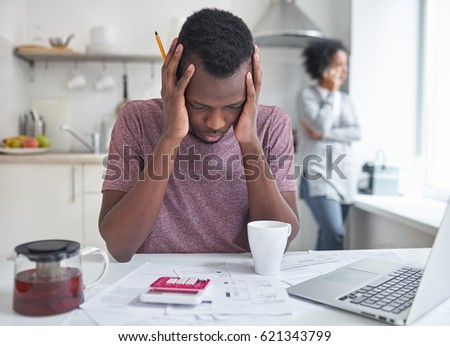 Stressed unemployed african american male having debts, holding head in despair, can not pay bills. His wife using mobile phone, talking with bank worker in background. Financial problems concept