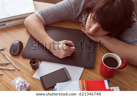 A girl draws a picture on a graphic tablet. She looks tiredly at the monitor. Various items on the table.