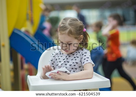 A girl of European appearance, 3-4 years old, sits on a chair and looks at the smartphone. A child is playing or watching a cartoon.