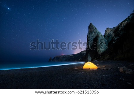 Glowing camping tent on a beautiful sea shore with rocks at night under a starry sky Royalty-Free Stock Photo #621513956