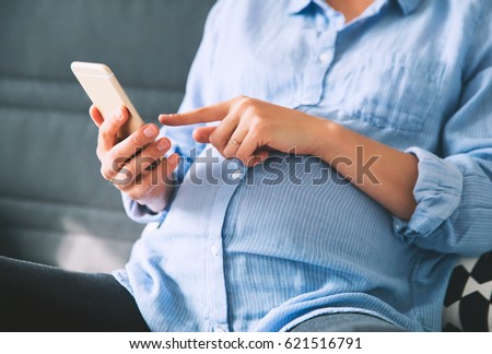 Close-up photo of pregnant woman holding and use mobile smart phone at home interiors. Pregnancy, Technology, preparation and expectation concept. Background with copy space.