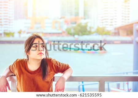 Beautiful biracial teen girl resting arms on railing by the riverside with sunny  boat and urban scene in background