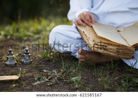 Boy in a kimono sits in a  yoga lotus position meditating, reading old open book of wisdom and there are a number  Tibetan bells  and burn incense at meadow Royalty-Free Stock Photo #621585167