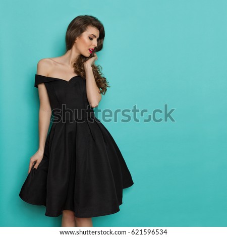 Beautiful young woman in elegant black cocktail dress is looking away. Three quarter length studio shot on turquoise background.