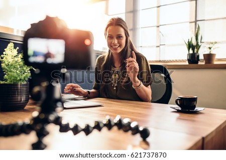 Young woman recording video for her vlog on a digital camera mounted on flexible tripod. Smiling woman sitting at her desk working on a laptop computer. Royalty-Free Stock Photo #621737870