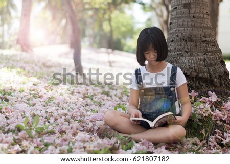 Adorable Asia female kid reading literacy in park. Child reader read young adult novel book in garden park with pink flowers o ground.