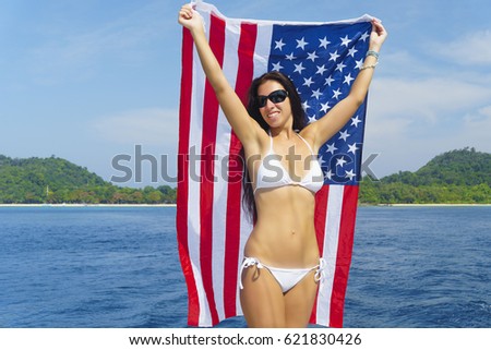 Beautiful brunette woman in white swimsuit is staying with usa flag on the stem of yacht at summer sunny day