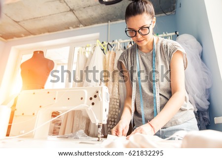 Young dressmaker woman sews clothes on sewing machine. Smiling seamstress and her hand close up in workshop. Focus on sewing machine and tissue. Royalty-Free Stock Photo #621832295