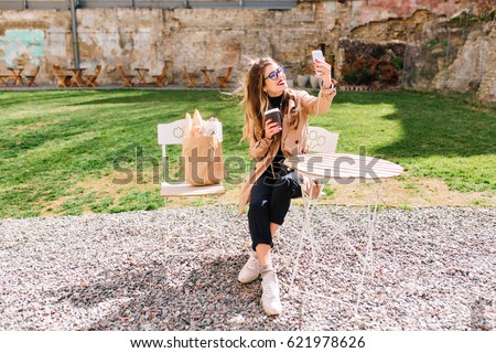 Attractive hipster girl wearing beige coat makes selfie fo  profile during coffee break in outdoor cafe. Young stylish woman taking pictures of herself in the park after food shopping