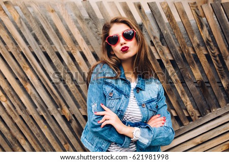 Kissing girl wearing heart glasses with long hair and arms folded on wooden background. Close-up portrait of lovely young woman with red lipstick in jeans jacket. Confident pose, body language Royalty-Free Stock Photo #621981998