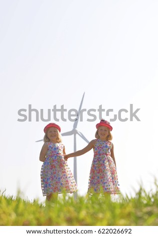 Twin sisters stand in front of a huge wind turbine and smile at the camera.
Their faces express happiness for this type of environmental energy.