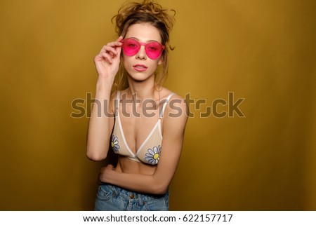 A portrait of a girl in an amusing swimsuit, jeans, with carelessly gathered hair. A girl stands holding on the shade of pink glasses and looks at the camera