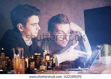 Two handsome young men using laptop and discussing new project at workplace with abstract night city view. Team work concept. Double exposure
