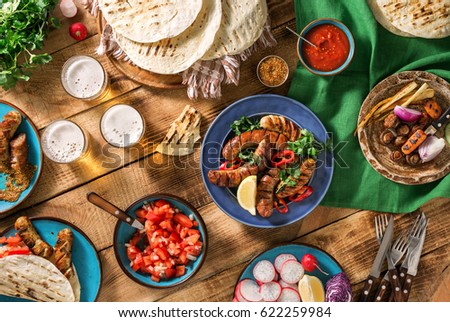 Dinner party food concept. Dinner table with grilled sausage, tortilla wraps, beer drink and different dishes on wooden table, rustic style