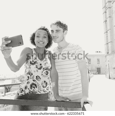 Black and white portrait of ethnically diverse couple sightseeing in destination city monument, holding smart phone taking selfies, holiday together outdoors. Travel technology lifestyle recreation.