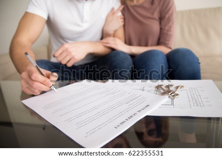 Close up of tenants signing rental agreement, renters couple sitting on couch, male hand with a pen putting signature, focus on document and keys, joint residential tenancy, lease contract 