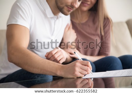 Close up of couple signing documents, young man putting signature on document, his wife sitting next to husband holding his arm, real estate purchase, first time home buyers, prenuptial agreement  Royalty-Free Stock Photo #622355582