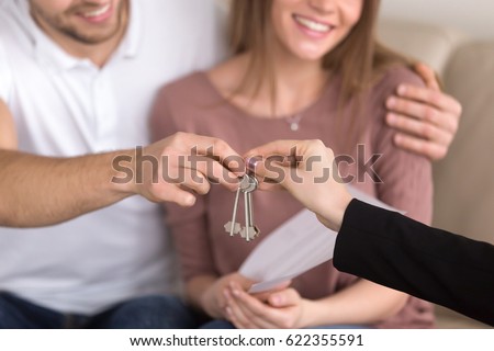 Close up of happy couple getting keys to their new house, hand of a female real estate agent giving apartment keys to a man sitting on sofa embracing his girlfriend, renting or buying home concept 