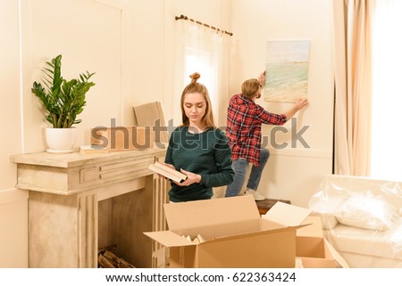 serious woman unpacking box and man hanging picture at new home