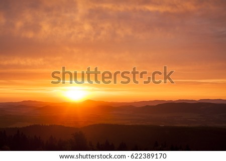 Natural Sunset Sunrise Scene Over Highland or Mountains. Bright Dramatic Sky And Dark Ground. Countryside Landscape Under Scenic Colorful Sky At Daybreak Dawn. Sun Over Skyline, Horizon. Warm Colors.
