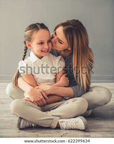 Beautiful woman and her cute little daughter are hugging and smiling while sitting on the floor, mom is kissing her daughter in cheek