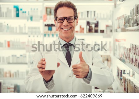 Handsome pharmacist is holding a medication, showing Ok sign and smiling while standing in pharmacy
