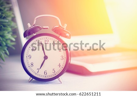 Old analog alarm clock and blurred laptop on white wooden table toned