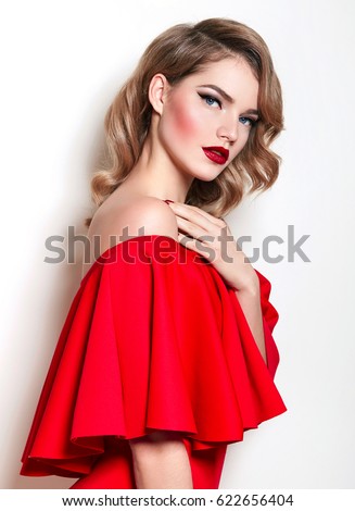 A beautiful slender young girl in a red dress and with red lips on a white background is standing. Retro hairstyle and makeup. Arrows, red lipstick. Advertising, fashion, beauty.