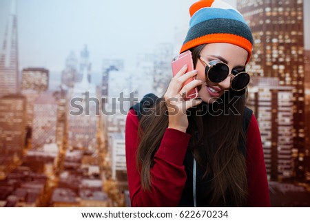 A young and positive girl with sunglasses is talking on the phone in her room against the background of the city depicted on the wallpaper.