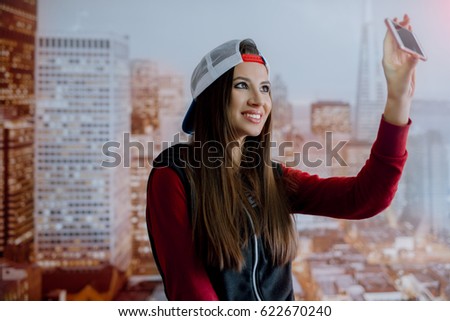 A young and positive girl is photographed on her smartphone in her room on the background of the city depicted on the wallpaper.