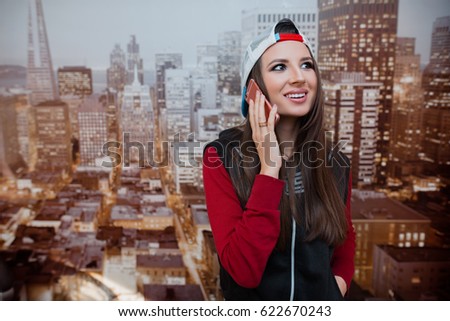 A young and positive girl is talking on the phone in her room against the backdrop of the city depicted on the wallpaper.