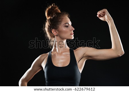 Photo of young sports woman standing over black background and showing her biceps. Looking aside.