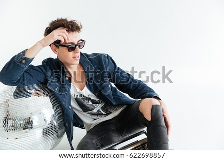 Picture of concentrated young man wearing sunglasses sitting isolated over white background near disco ball and boombox. Looking aside.