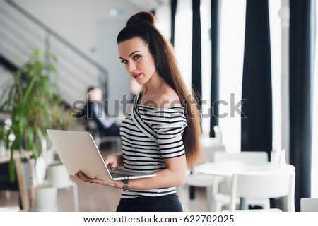Beautiful young business woman with long hair, keeps laptop in hand, smiling, looking at camera.