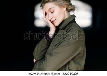 Lonely beautiful and attractive young blonde girl in green coat standing in a dark tunnel with abstract background  and dreaming with closed eyes. Pensive, thoughtful. Contemplation and mindfulness.